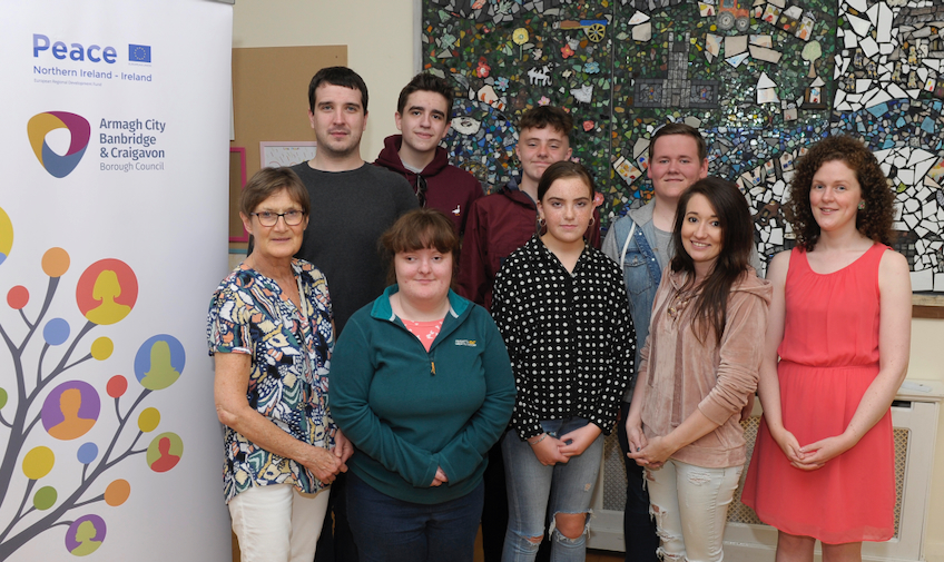 Photo of a smiling group of teenagers and adults, standing in front of a mural, with an Armagh City. Banbridge & Craigavon pop-up display, with a Peace Northern Ireland logo and a graphic of a tree with outlines of people's heads in circular 'leaves'