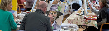 Photo of a group of adults seated at tables with easels in front of them, painting portraits.